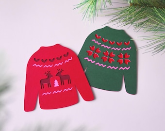 May include: Two paper Christmas sweater cutouts. One is red with reindeer and a tree, the other is green with snowflake and heart designs. Both have pink and white accents. Festive holiday decor.