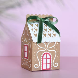 Könnte beinhalten: Eine Geschenkbox in Form eines Lebkuchenhauses aus braunem Karton, verziert mit weißen Zuckergussdetails und einem roten Fenster. Das Dach hat ein weißes, herzförmiges Ausschnittmuster und ist mit einem grünen Samtband verziert. Ideal für Weihnachtsgeschenke.