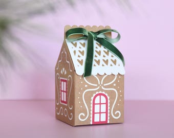 May include: A gingerbread house-shaped gift box made of brown cardboard, decorated with white icing details and a red window. The roof has a white, heart-shaped cutout design and is topped with a green velvet ribbon. Perfect for holiday gifting.