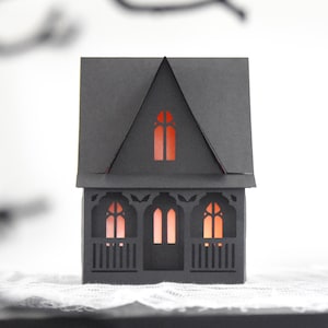 May include: A black paper haunted house with orange glowing windows and a detailed porch. The house has a triangular roof and is designed for Halloween decorations. The house is sitting on a white surface.