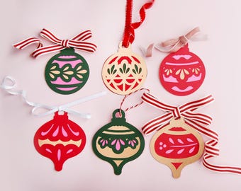 May include: A collection of colorful Christmas ornaments with decorative cut-out designs. The ornaments are in various shapes, including round and teardrop, and feature red, green, and gold colors. Each ornament is adorned with a ribbon or string for hanging.