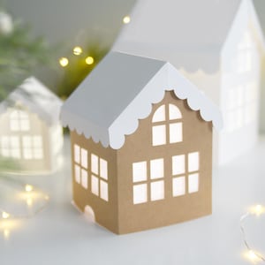 May include: A small house-shaped paper lantern with brown walls and a white scalloped roof. The structure features cut-out windows that emit a soft light, creating a glowing effect. The design mimics a gingerbread house with a peaked roof.