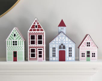 May include: A set of four decorative paper houses in varying sizes and colors. The houses feature detailed architectural patterns including windows, doors, and roof designs in shades of green, red, light blue, and pink with white accents.