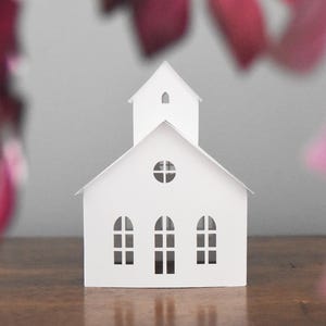 Puede incluir: Maqueta de iglesia de papel blanco con campanario y ventanas arqueadas. El modelo está sobre una superficie de madera, con elementos borrosos rosas y rojos en primer plano.