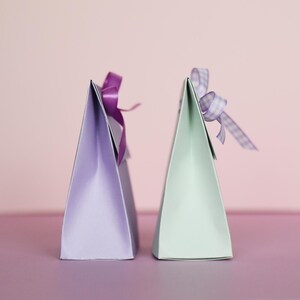 May include: Two triangular gift boxes, one lavender and one mint green, stand side-by-side. Each box is adorned with a matching ribbon bow. The boxes are on a purple surface, with a soft pink background.