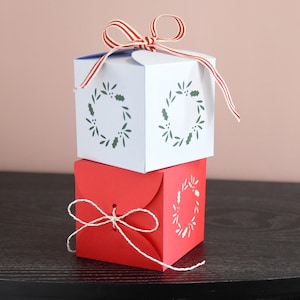Könnte beinhalten: Zwei gestapelte Geschenkboxen. Die obere Box ist weiß mit einem rot-weiß gestreiften Band und einem Stechpalmenkranz-Design. Die untere Box ist rot mit einer weißen Schnurschleife und einem passenden Stechpalmenkranz-Design.