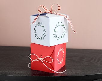 May include: Two stacked gift boxes. The top box is white with a red and white striped ribbon and a holly wreath design. The bottom box is red with a white string bow and a matching holly wreath design.