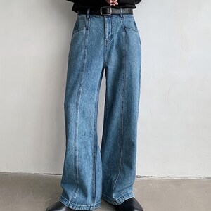 K&ouml;nnte beinhalten: Hellblaue Jeans mit weitem Bein. Die Jeans haben einen klassischen F&uuml;nf-Taschen-Stil, einen schwarzen G&uuml;rtel und vertikale Nahtdetails auf der Vorderseite jedes Beins. Die Jeans werden mit schwarzen Schuhen getragen.