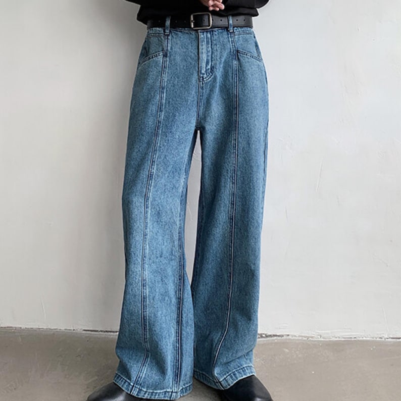 K&ouml;nnte beinhalten: Hellblaue Jeans mit weitem Bein. Die Jeans haben einen klassischen F&uuml;nf-Taschen-Stil, einen schwarzen G&uuml;rtel und vertikale Nahtdetails auf der Vorderseite jedes Beins. Die Jeans werden mit schwarzen Schuhen getragen.