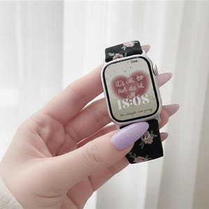 Puede incluir: Un reloj inteligente plateado con una correa de tela negra con un peque&ntilde;o estampado floral rosa y verde. La esfera muestra un gr&aacute;fico de coraz&oacute;n rojo con el texto "it's ok to not do it all" y la hora "18:08" en n&uacute;meros digitales blancos.