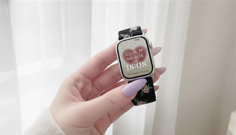 Puede incluir: Un reloj inteligente plateado con una correa de tela negra con un peque&ntilde;o estampado floral rosa y verde. La esfera muestra un gr&aacute;fico de coraz&oacute;n rojo con el texto "it's ok to not do it all" y la hora "18:08" en n&uacute;meros digitales blancos.