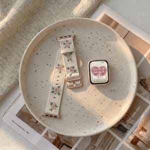 Puede incluir: Un reloj inteligente con marco en oro rosa y pantalla digital que muestra el texto 'it's ok to not do it all' y '18:08', junto a una correa de tela color crema con un patr&oacute;n bordado de peque&ntilde;as rosas rosadas y hojas verdes.