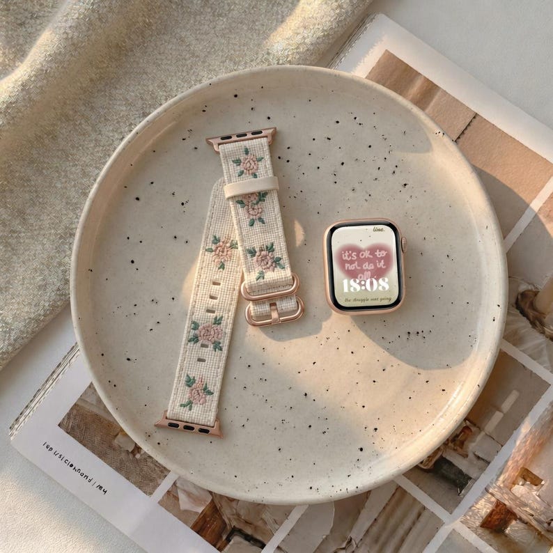 Puede incluir: Un reloj inteligente con marco en oro rosa y pantalla digital que muestra el texto 'it's ok to not do it all' y '18:08', junto a una correa de tela color crema con un patr&oacute;n bordado de peque&ntilde;as rosas rosadas y hojas verdes.