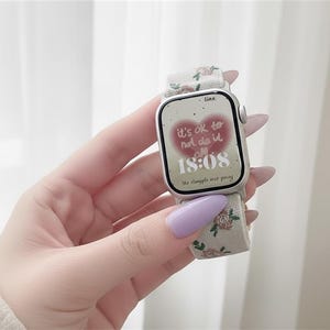 Puede incluir: Un reloj inteligente plateado con una correa de tela color crema con un peque&ntilde;o estampado floral. La pantalla digital muestra un gr&aacute;fico de coraz&oacute;n rojo con el texto 'it's ok to not do it all' y la hora '18:08' en fuente blanca.