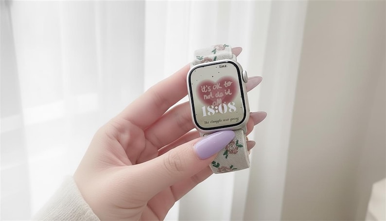 Puede incluir: Un reloj inteligente plateado con una correa de tela color crema con un peque&ntilde;o estampado floral. La pantalla digital muestra un gr&aacute;fico de coraz&oacute;n rojo con el texto 'it's ok to not do it all' y la hora '18:08' en fuente blanca.