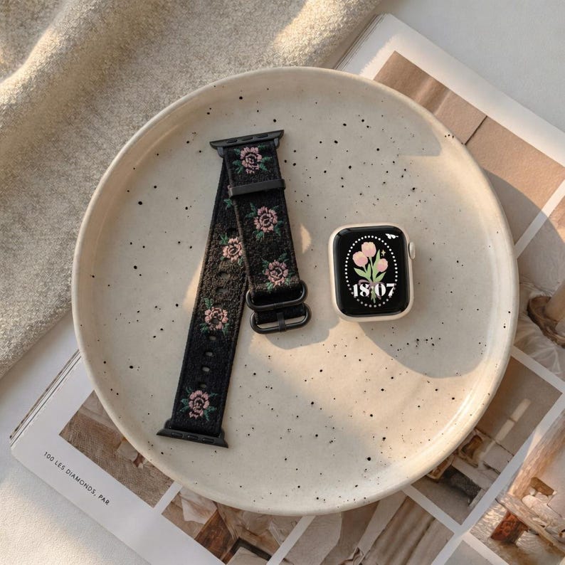 Puede incluir: Una correa de reloj inteligente negra con un patr&oacute;n floral bordado en hilo rosa y verde, junto a un reloj inteligente que muestra una esfera digital con la hora 18:07 y un gr&aacute;fico de tulipanes rosas sobre fondo negro con borde blanco.