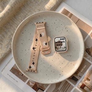 Puede incluir: Una correa de reloj tejida en color beige con peque&ntilde;os bordados florales en negro y blanco y una caja de reloj inteligente a juego. La esfera muestra el texto AND wish you every day 17:39 en una fuente manuscrita sobre fondo claro.