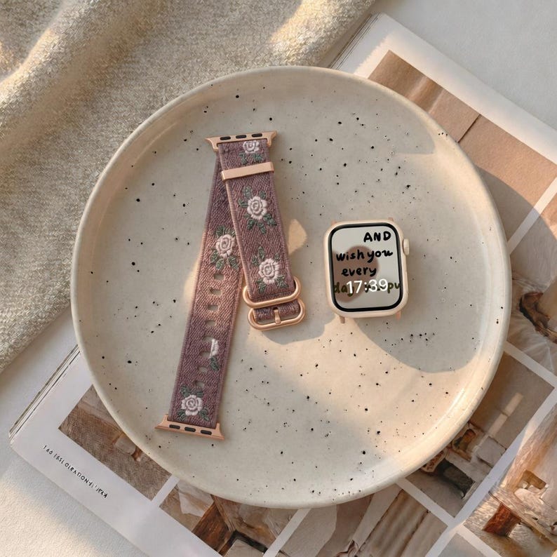 Puede incluir: Una correa de reloj malva con un patr&oacute;n de rosas blancas y herrajes en oro rosa, junto a una caja de reloj a juego de color claro que muestra el texto AND wish you every day 17:39 en la pantalla, sobre un plato color crema moteado.