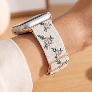 Puede incluir: Una correa para reloj inteligente de tela color blanco hueso con un patr&oacute;n bordado de peque&ntilde;as rosas rosadas y hojas verdes. La correa incluye herrajes met&aacute;licos en color oro rosa, con una hebilla deslizante ajustable, unida a una caja plateada.