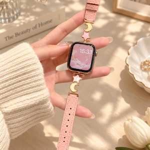 Pode incluir: Um smartwatch com moldura em ouro rosa e pulseira de couro texturizado rosa. A pulseira apresenta berloques de metal dourado em formato de luas crescentes e estrelas. O mostrador exibe a hora 15:33 sobre um fundo rosa com nuvens.