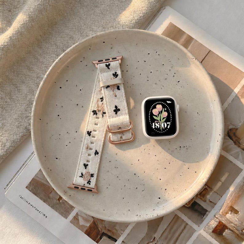 Puede incluir: Un reloj inteligente con caja blanca y pantalla digital que muestra un gr&aacute;fico de tulipanes rosas y la hora 18:07, junto a una correa de tela color crema con peque&ntilde;os bordados de flores negras y rosas y herrajes en tono oro rosa.