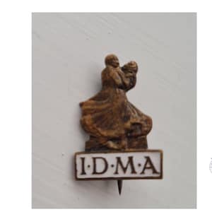 May include: A brown, sculpted metal pin featuring two figures in a dancing pose above a rectangular white base. The base contains the black text I.D.M.A. in capital letters, separated by dots. The pin has a pointed metal fastener at the bottom.