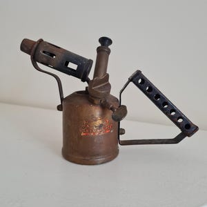 May include: A vintage brass blowtorch featuring a cylindrical fuel tank with a weathered patina, a metal burner nozzle, and a triangular handle with circular cutouts. The tank displays a faded logo with the text "Primus" and "MADE IN SWEDEN".