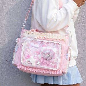 Puede incluir: Bolso de hombro rosa con un bolsillo frontal transparente que muestra un personaje de dibujos animados, nubes y estrellas. El bolso presenta volantes, un lazo y un charm en forma de estrella. Tiene una correa rosa y un bolsillo lateral con cremallera.