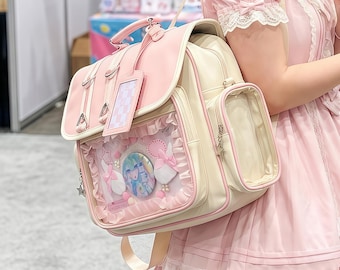 May include: A pink and cream-colored backpack with a clear front pocket displaying decorative elements. The bag features a top handle, shoulder straps, and a small side pocket. Accents include a pink tag and silver hardware.