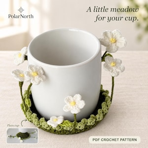 May include: A white ceramic mug sits inside a green crocheted coaster featuring white flowers with yellow centers. The text reads PolarNorth, A little meadow for your cup, Place cup, and PDF CROCHET PATTERN. The coaster has a circular base.