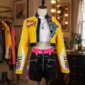 May include: A yellow cropped jacket with black trim, a white top, and black shorts. The jacket features a Rrrrrrumi patch, a silver chain necklace, and colorful graphic patterns on the sleeves. The black shorts have a front zipper and pink belt loops.