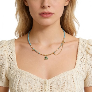 Puede incluir: Un collar de cuentas turquesas con detalles dorados y un colgante triangular. El collar presenta pequeñas cuentas turquesas, detalles metálicos dorados y una delicada cadena. El colgante tiene un centro verde y un marco dorado.