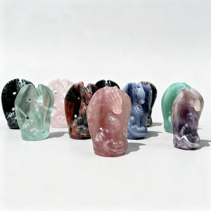 Könnte beinhalten: Eine Sammlung von geschnitzten Drachenfiguren aus Edelsteinen in verschiedenen Farben, darunter Grün, Rosa, Rot, Blau und Lila. Die Drachenskulpturen werden auf einer weißen Oberfläche ausgestellt. Die Schnitzereien weisen detaillierte Merkmale auf.