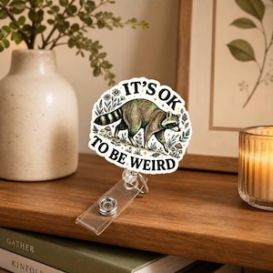 Puede incluir: Un portatarjetas circular con una ilustración de un mapache rodeado de flores y setas. El texto IT'S OK TO BE WEIRD está impreso en letras mayúsculas negras alrededor del mapache. El accesorio incluye una correa de plástico transparente.