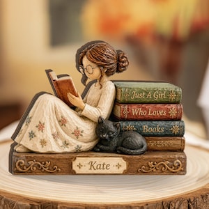 Puede incluir: Una figura de resina de una persona con el pelo castaño recogido, gafas y vestido floral, leyendo un libro. Un gato negro descansa junto a una pila de cuatro libros con el texto "Just A Girl Who Loves Books". La base está grabada con "Kate".