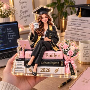 Peut inclure: Un présentoir en acrylique lumineux représentant un diplômé en toge noire, tenant une tasse marquée Future Engineer et un livre intitulé Class of 2026. La figurine est assise sur une pile de livres marqués Emily, Software Engineer et 2026.