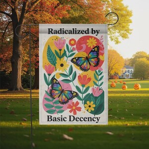 Peut inclure: Un drapeau de jardin avec le texte "Radicalized by Basic Decency" et un motif floral et papillon coloré. Le drapeau présente des éléments roses, jaunes et verts, avec deux papillons et diverses fleurs. Le drapeau est affiché à l'extérieur.