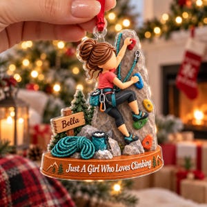 May include: A clear acrylic Christmas ornament featuring a climber on a rock wall with colorful holds. The figure has brown hair in a bun, wearing a red shirt and black leggings. It includes a sign labeled Bella, a rope, and the text Just A Girl Who Loves Climbing.