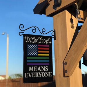 May include: Black sign with the text "We the People MEANS EVERYONE" and a flag design combining the American flag with the rainbow pride flag. The sign is hanging from a decorative black metal bracket.