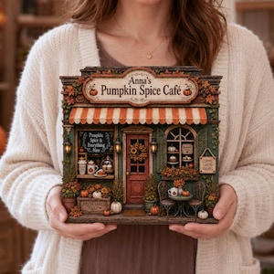 Op de afbeelding: Een miniatuurmodel van een café genaamd Anna's Pumpkin Spice Café, met een oranje-wit gestreepte luifel, een rode deur met een krans en etalages met pompoenen, koffiemokken en bordjes met de tekst Pumpkin Spice & Everything Nice.
