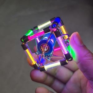 May include: A geometric, illuminated cube with colorful LED lights. The cube is held in a hand and features a central component with a blue cylinder and circuit board. The lights include pink, green, yellow, and white.