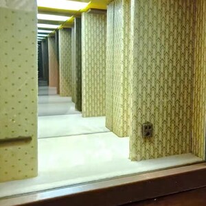 Może przedstawiać: Miniaturowa diorama długiego korytarza z ż&oacute;łtym oświetleniem sufitowym, ścianami pokrytymi powtarzającym się zielono-białym wzorem geometrycznym oraz małym gniazdkiem elektrycznym zamontowanym na ścianie. Scena tworzy iluzję głębi.