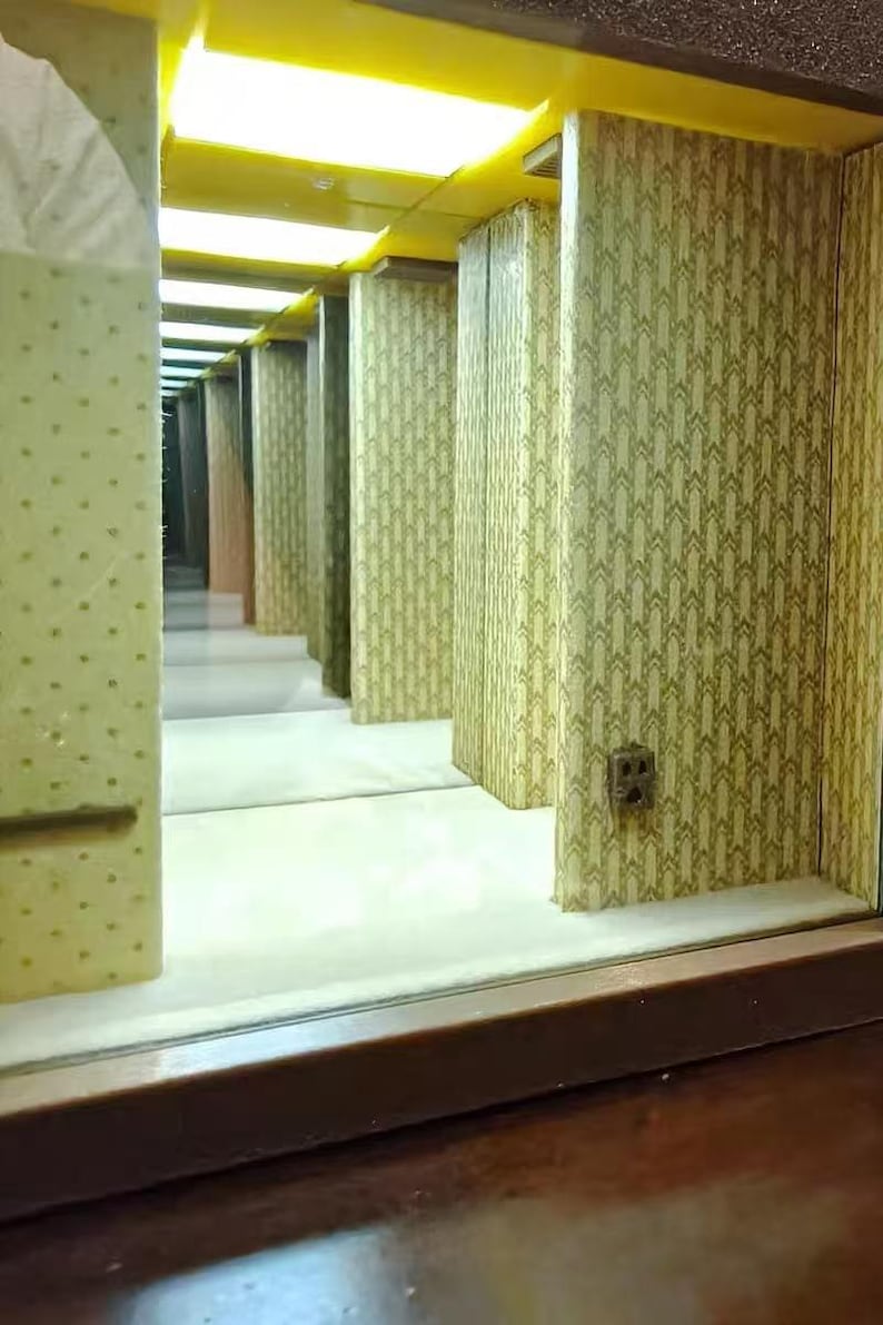 Może przedstawiać: Miniaturowa diorama długiego korytarza z ż&oacute;łtym oświetleniem sufitowym, ścianami pokrytymi powtarzającym się zielono-białym wzorem geometrycznym oraz małym gniazdkiem elektrycznym zamontowanym na ścianie. Scena tworzy iluzję głębi.