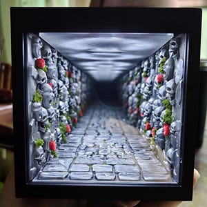 Könnte beinhalten: Ein schwarz gerahmtes Diorama mit einem tiefen, beleuchteten Korridor, einem Boden aus Steinoptik und Wänden, die mit grauen Schädeln, kleinen grünen Moosflecken und winzigen roten Blumen besetzt sind, was die Illusion eines langen Tunnels erzeugt.