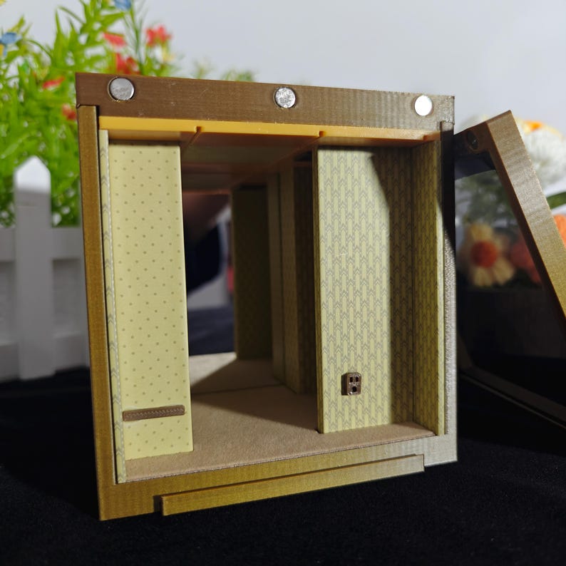Może przedstawiać: Miniaturowy pok&oacute;j w pudełku z brązową ramą, z ż&oacute;łtymi ścianami o subtelnym wzorze w kropki po lewej stronie i geometrycznym wzorem w strzałki po prawej. Wnętrze zawiera mały detal w postaci brązowego gniazdka elektrycznego na ścianie.