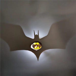 K&ouml;nnte beinhalten: Eine Batman-Symbol-Leuchte, die einen Schatten des ikonischen fledermausf&ouml;rmigen Emblems wirft. Das Licht ist gelb und die Leuchte ist braun. Der Schatten ist gro&szlig; und vor einem grauen Hintergrund prominent. Das Thema ist Superhelden-Dekor.