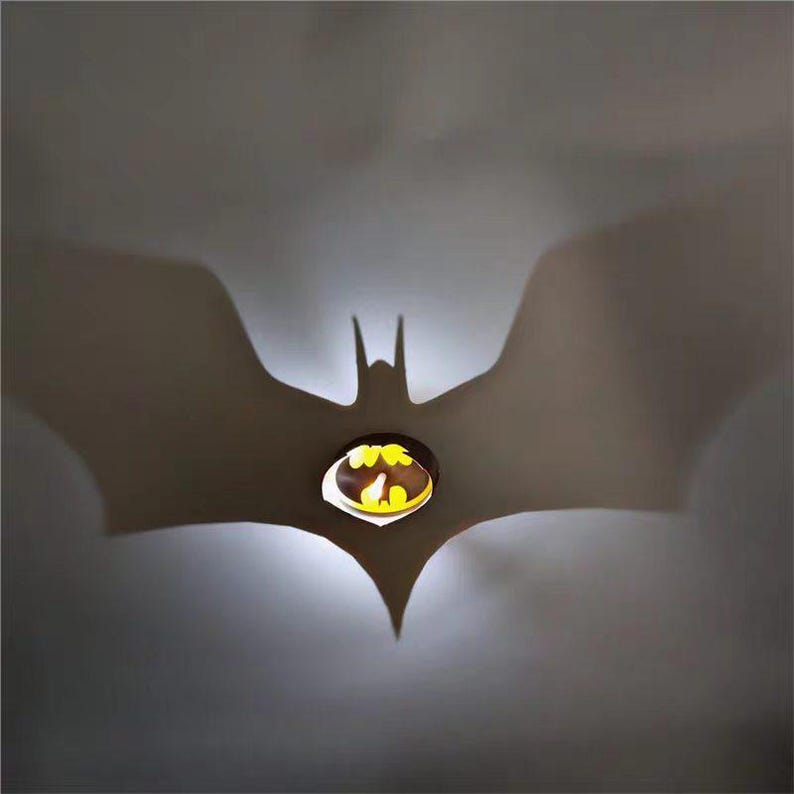 K&ouml;nnte beinhalten: Eine Batman-Symbol-Leuchte, die einen Schatten des ikonischen fledermausf&ouml;rmigen Emblems wirft. Das Licht ist gelb und die Leuchte ist braun. Der Schatten ist gro&szlig; und vor einem grauen Hintergrund prominent. Das Thema ist Superhelden-Dekor.