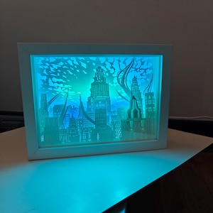 Può includere: Un'opera d'arte in carta incorniciata che raffigura un paesaggio urbano futuristico. L'opera presenta ritagli di carta a strati di edifici, illuminati con luci a LED blu e verdi. La cornice bianca circonda il paesaggio urbano dettagliato.