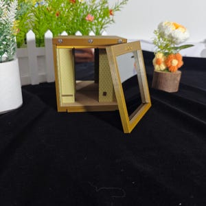 Może przedstawiać: Miniaturowe drewniane pudełko z ramą w kolorze złota i otwieranym przednim panelem. Wnętrze ma jasne ż&oacute;łte ściany z subtelnym wzorem w kropki, małą p&oacute;łkę i teksturowaną podłogę. Konstrukcja jest otwarta z przezroczystym przednim oknem.