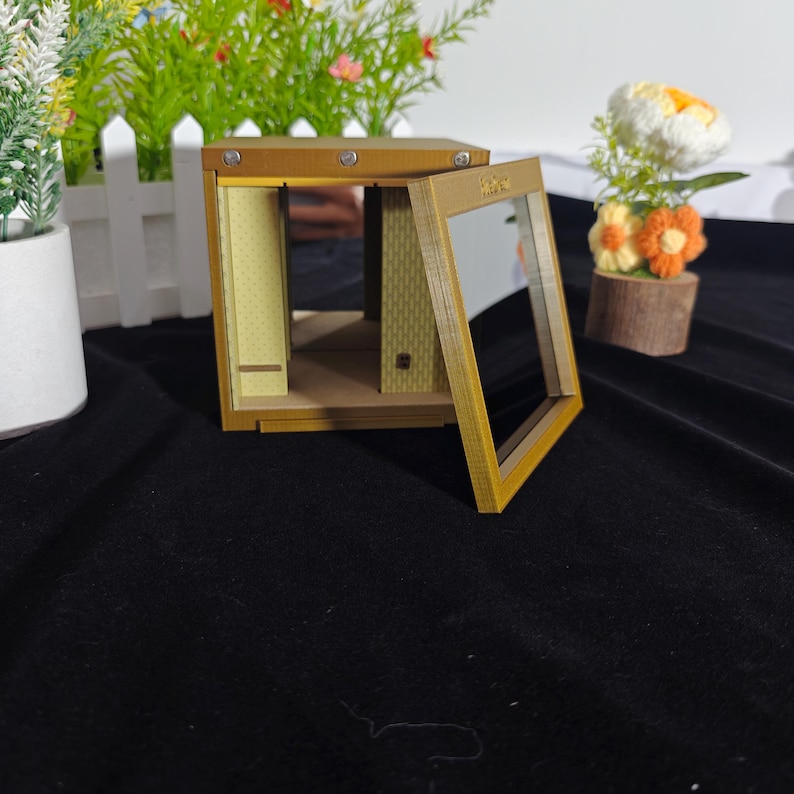 Może przedstawiać: Miniaturowe drewniane pudełko z ramą w kolorze złota i otwieranym przednim panelem. Wnętrze ma jasne ż&oacute;łte ściany z subtelnym wzorem w kropki, małą p&oacute;łkę i teksturowaną podłogę. Konstrukcja jest otwarta z przezroczystym przednim oknem.